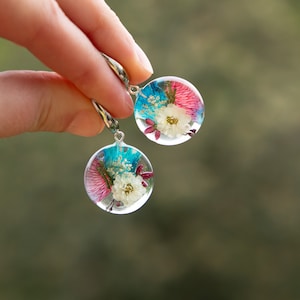 May include: Clear, round earrings with colorful dried flowers encased in resin. The earrings feature a white flower, pink and blue accents, and silver-colored hooks. The earrings are held up by a hand.