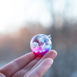 May include: A small glass sphere pendant with a pink, blue, and white gradient. The sphere contains a white cloud-like design with a hint of sparkle.