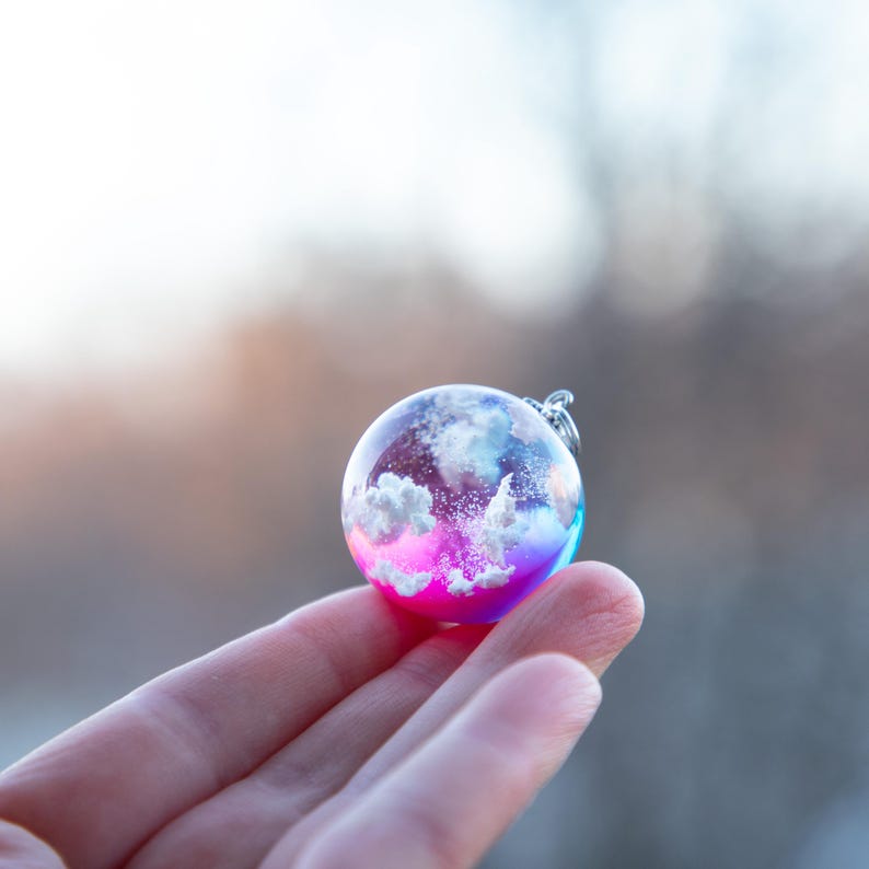 May include: A small glass sphere pendant with a pink, blue, and white gradient. The sphere contains a white cloud-like design with a hint of sparkle.