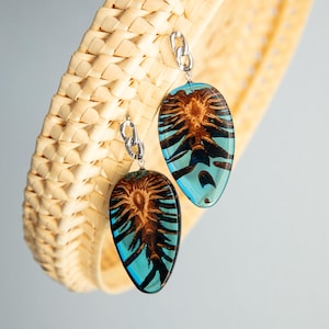 May include: A pair of teardrop-shaped earrings with a silver chain and a blue resin setting. The resin contains a dried pine cone.