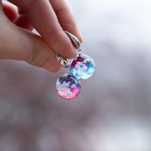 May include: A pair of silver hoop earrings with round, colorful resin charms. The charms feature white clouds against a blue and pink gradient background.