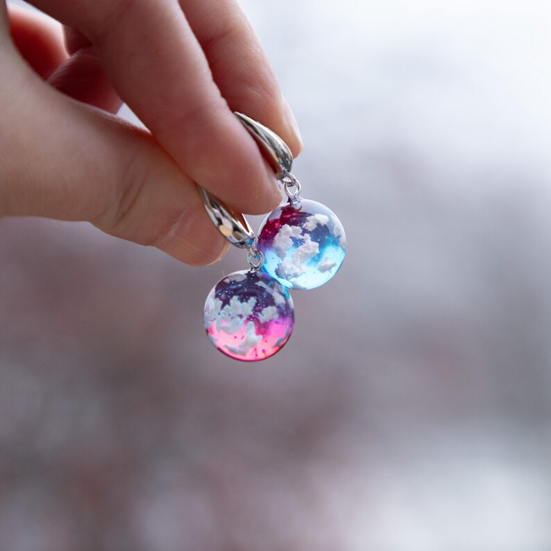May include: A pair of silver hoop earrings with round, colorful resin charms. The charms feature white clouds against a blue and pink gradient background.