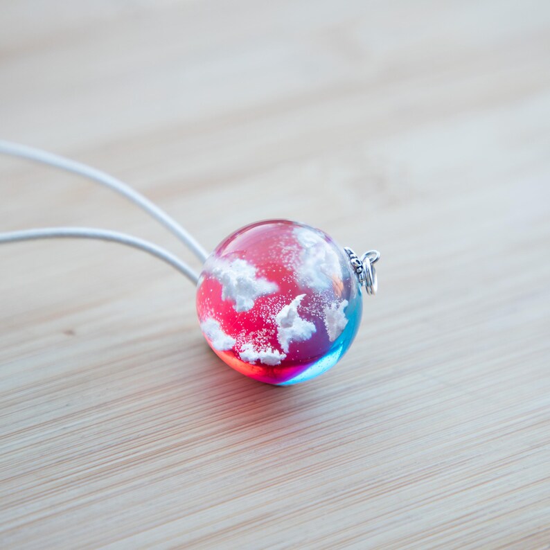 May include: A round glass pendant necklace with a red and blue resin background and white clouds inside. The pendant is attached to a white cord.