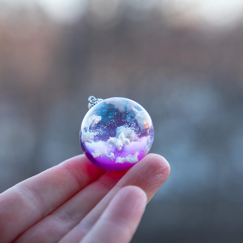 May include: A small glass sphere pendant with a white cloud design inside. The sphere is filled with a purple and blue resin, creating a cloudy sky effect.
