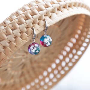 May include: A pair of silver hoop earrings with round, colorful resin dangles. The dangles feature a swirling pattern of blue, pink, and white.