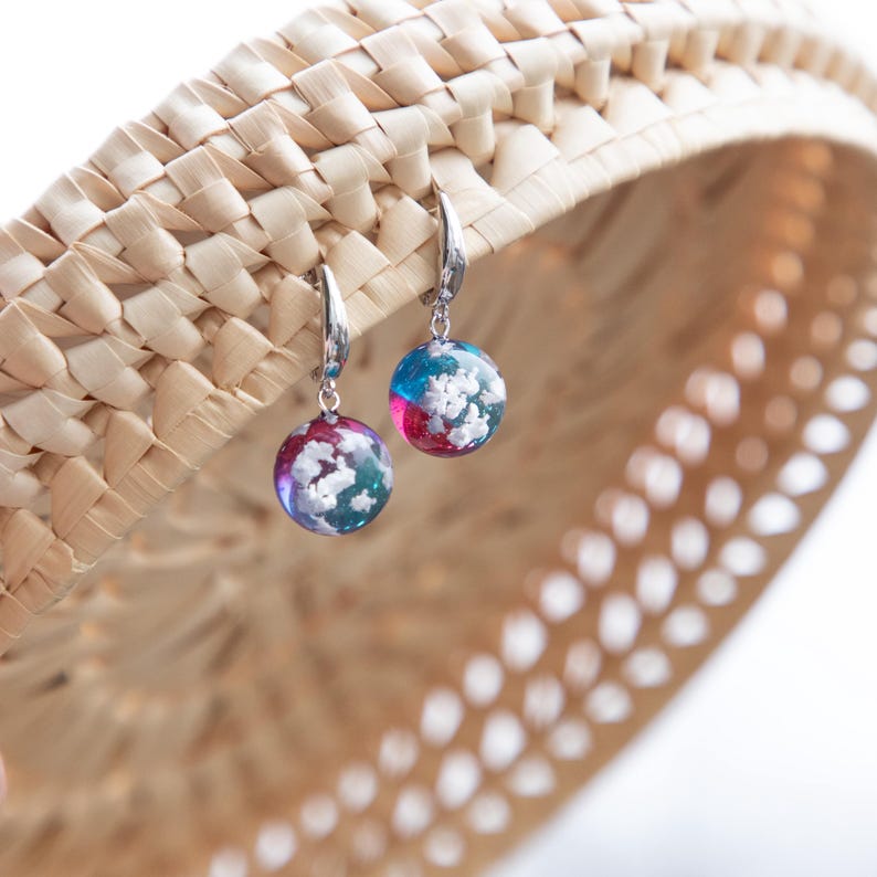 May include: A pair of silver hoop earrings with round, colorful resin dangles. The dangles feature a swirling pattern of blue, pink, and white.