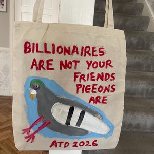 Op de afbeelding: Een beige canvas draagtas met een handgeschilderde illustratie van een grijze duif met een groene kop en rode poten. De tekst luidt: BILLIONAIRES ARE NOT YOUR FRIENDS PIGEONS ARE, met ATD 2026 onderaan in rode verf gedrukt.
