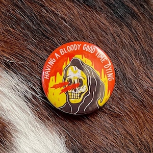 May include: A red, yellow, and black button with a cartoon image of the Grim Reaper with flames behind it. The text on the button reads "Having a bloody good time dying."
