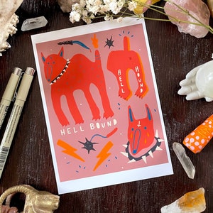 May include: A pink and red illustration of a dog with the text "Hell Hound" and "Hell Bound". The dog is depicted with a spiked collar and a cross on its forehead.