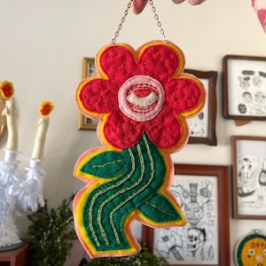 Könnte beinhalten: Ein Wandbehang aus Filz in Form einer Blume mit einer roten Blüte, grünen Blättern und einem gelben Stiel. Die Blüte hat ein weißes Auge mit einem roten Zentrum.