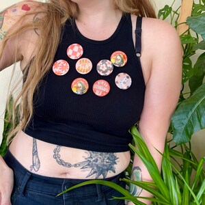 K&ouml;nnte beinhalten: Eine Person tr&auml;gt ein schwarzes Tanktop mit acht bunten Anstecknadeln. Die Nadeln zeigen verschiedene Designs, darunter Blumen, einen Totenkopf und einen Text, der "You're Outshining Me" lautet.