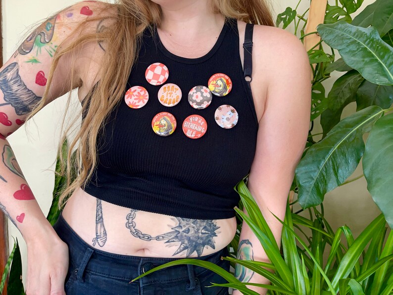 K&ouml;nnte beinhalten: Eine Person tr&auml;gt ein schwarzes Tanktop mit acht bunten Anstecknadeln. Die Nadeln zeigen verschiedene Designs, darunter Blumen, einen Totenkopf und einen Text, der "You're Outshining Me" lautet.