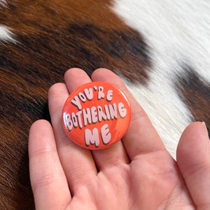 K&ouml;nnte beinhalten: Ein runder orangefarbener Knopf mit dem Text "You're Bothering Me" in rosa Buchstaben.