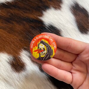 May include: A round button with a black and white illustration of a skeleton wearing a hooded robe. The skeleton has red eyes with flames coming out of them. The text around the skeleton reads "Having a bloody good time dying".