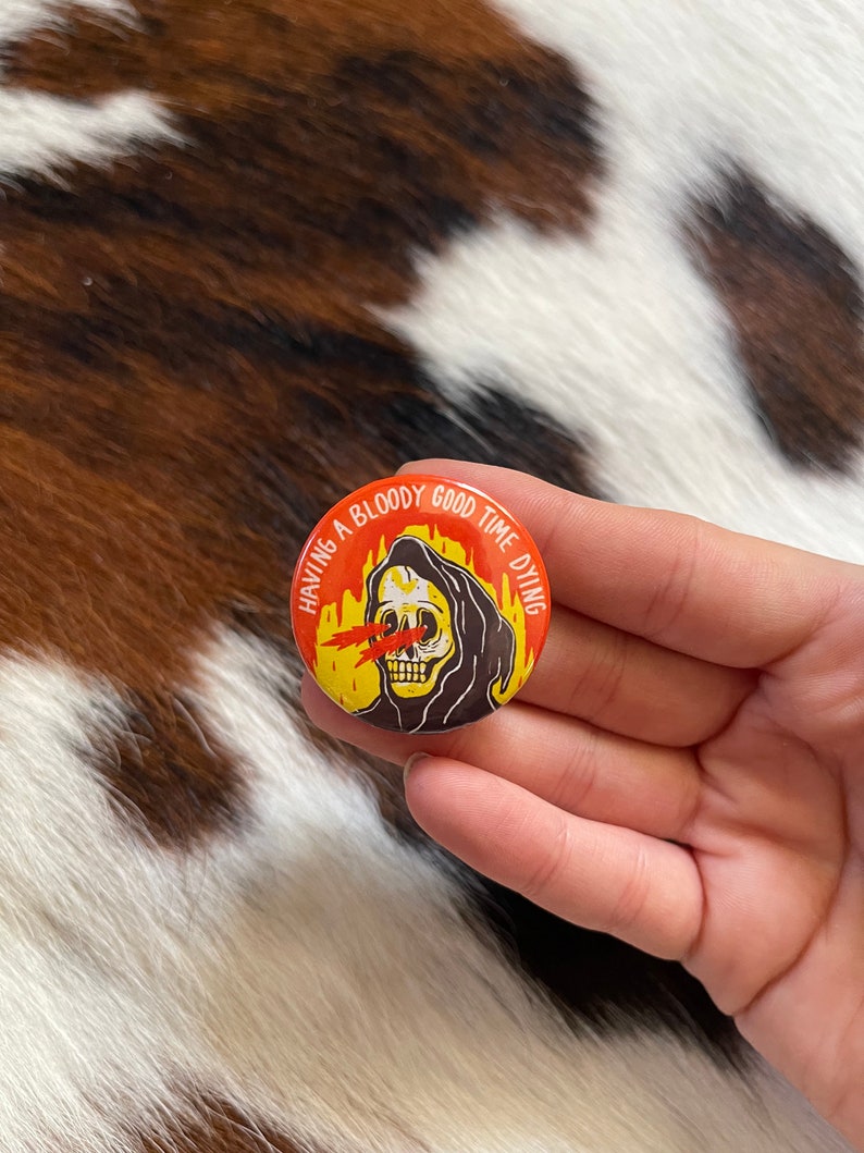 May include: A round button with a black and white illustration of a skeleton wearing a hooded robe. The skeleton has red eyes with flames coming out of them. The text around the skeleton reads "Having a bloody good time dying".