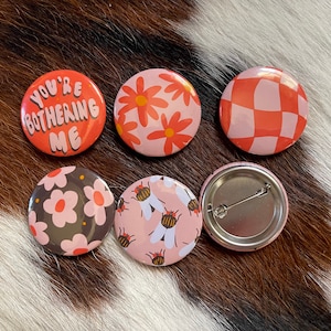 K&ouml;nnte beinhalten: Sechs runde Anstecknadeln mit verschiedenen Designs. Eine Nadel sagt "You're Bothering Me" in rosa und wei&szlig;en Buchstaben auf rotem Hintergrund. Eine weitere Nadel hat ein rosa-wei&szlig;es Karomuster. Zwei Nadeln haben ein rosa-wei&szlig;es Blumenmuster. Eine Nadel hat ein rosa-wei&szlig;es Muster mit Bienen. Eine Nadel ist leer mit einer silbernen Nadelr&uuml;ckseite.