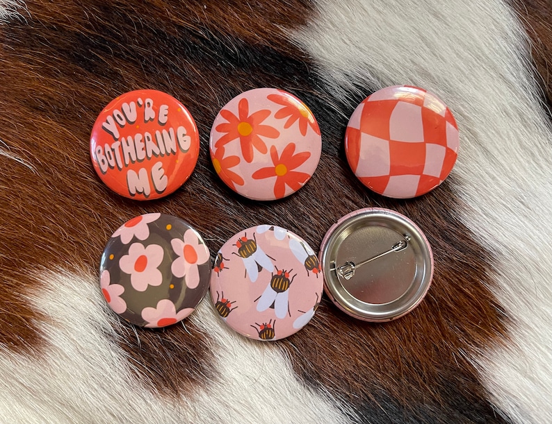 K&ouml;nnte beinhalten: Sechs runde Anstecknadeln mit verschiedenen Designs. Eine Nadel sagt "You're Bothering Me" in rosa und wei&szlig;en Buchstaben auf rotem Hintergrund. Eine weitere Nadel hat ein rosa-wei&szlig;es Karomuster. Zwei Nadeln haben ein rosa-wei&szlig;es Blumenmuster. Eine Nadel hat ein rosa-wei&szlig;es Muster mit Bienen. Eine Nadel ist leer mit einer silbernen Nadelr&uuml;ckseite.