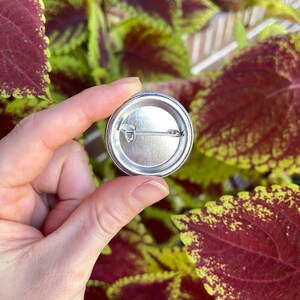 May include: The back of a silver button with a pin attached. The button is round and has a smooth surface.