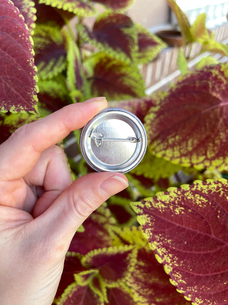 May include: The back of a silver button with a pin attached. The button is round and has a smooth surface.
