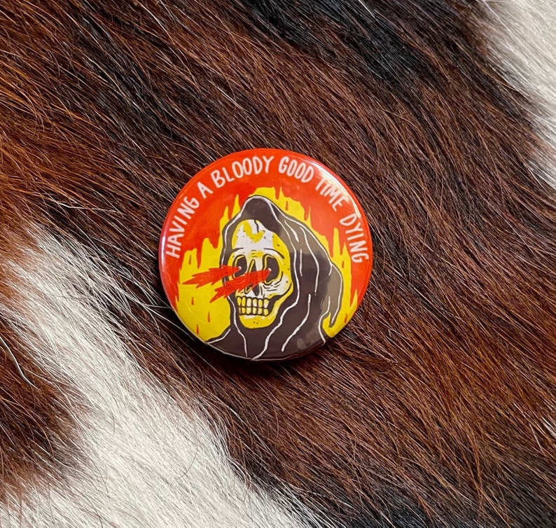 May include: A red, yellow, and black button with a cartoon image of the Grim Reaper with flames behind it. The text on the button reads "Having a bloody good time dying."