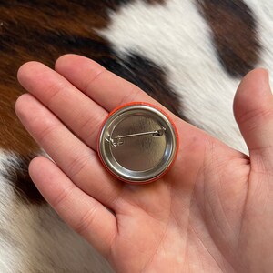 May include: The back of a round orange button with a silver pin and a metal backing.