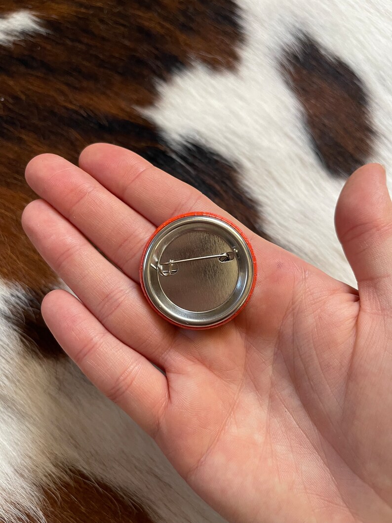 May include: The back of a round orange button with a silver pin and a metal backing.