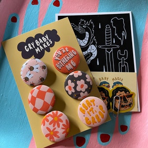 K&ouml;nnte beinhalten: Ein Set mit sechs bunten Anstecknadeln mit verschiedenen Designs, darunter Bienen, Blumen und karierte Muster. Die Nadeln werden auf einer gelben Karte mit dem Text "Cry Baby Makes" und "You're Bothering Me" angezeigt. Es gibt auch einen schwarz-wei&szlig;en Aufkleber mit einem Schwert- und Schlangenmotiv.