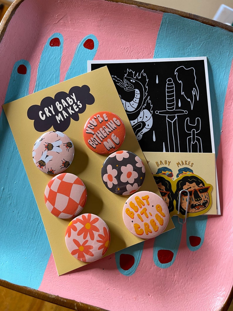 K&ouml;nnte beinhalten: Ein Set mit sechs bunten Anstecknadeln mit verschiedenen Designs, darunter Bienen, Blumen und karierte Muster. Die Nadeln werden auf einer gelben Karte mit dem Text "Cry Baby Makes" und "You're Bothering Me" angezeigt. Es gibt auch einen schwarz-wei&szlig;en Aufkleber mit einem Schwert- und Schlangenmotiv.