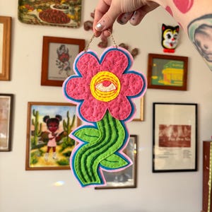 Può includere: Un'applique da parete a forma di fiore in feltro fatto a mano. Il fiore rosa ha un contorno blu e un centro giallo con un disegno a occhio. Lo stelo verde ha un design ondulato. Una catena dorata è attaccata per appenderlo.