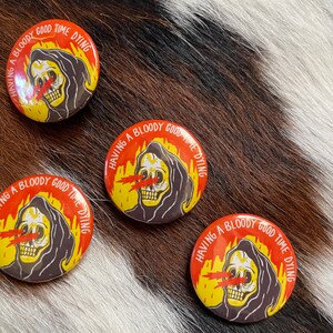 May include: Three round buttons with a black and white illustration of a skeleton wearing a hooded robe. The skeleton has red eyes and is surrounded by flames. The text on the buttons reads "Having a bloody good time dying."