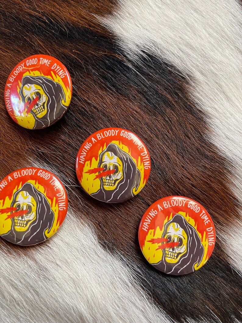 May include: Three round buttons with a black and white illustration of a skeleton wearing a hooded robe. The skeleton has red eyes and is surrounded by flames. The text on the buttons reads "Having a bloody good time dying."