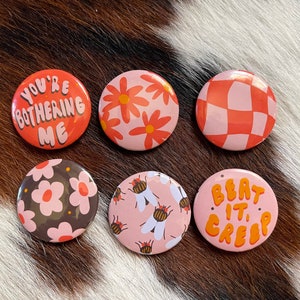 K&ouml;nnte beinhalten: Sechs runde rosa und orangefarbene Anstecknadeln mit verschiedenen Designs. Eine Nadel sagt "You're Bothering Me", eine andere hat ein kariertes Muster, eine andere hat ein Blumenmuster, eine andere hat ein Blumenmuster mit dunklem Hintergrund, eine andere hat ein Blumenmuster mit Bienen und eine andere sagt "Beat It Creep".