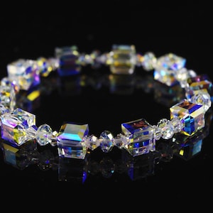 Puede incluir: Un brazalete de cristal con cuentas cuadradas iridiscentes y cuentas transparentes más pequeñas. El brazalete está sobre un fondo negro.