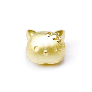 May include: A gold-coloured charm in the shape of Hello Kitty's head. The charm has a shiny, metallic finish.