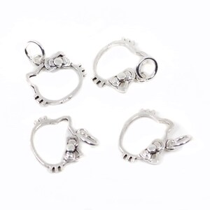 May include: Four silver-toned charms in the shape of a cat's head with a bow. Each charm has a small loop at the top for attaching to a necklace or bracelet. The charms are on a white background.