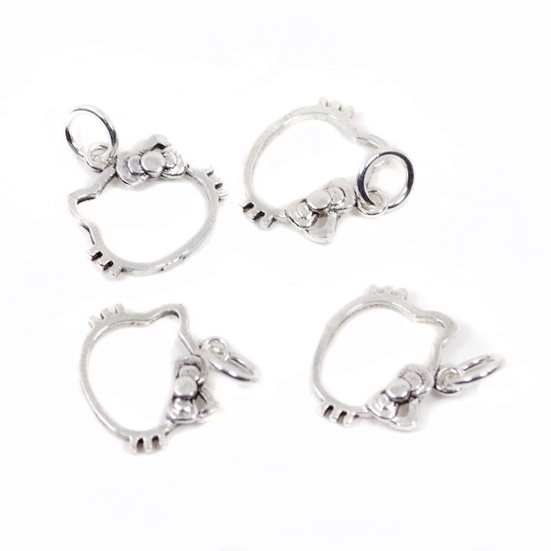 May include: Four silver-toned charms in the shape of a cat's head with a bow. Each charm has a small loop at the top for attaching to a necklace or bracelet. The charms are on a white background.