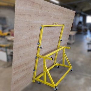 May include: A large, light brown wood-grain panel is supported by a bright yellow metal rolling cart. The cart features a sturdy A-frame design with multiple small wheels along the vertical supports and four swivel casters at the base.