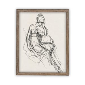 May include: A framed wall art piece featuring a charcoal sketch of a seated figure on a light cream background. The artwork uses loose, expressive black lines to define the form of the person, enclosed in a simple, textured medium-brown wooden frame.