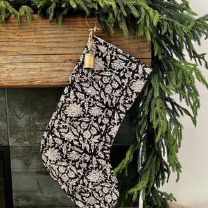 May include: A black and white floral patterned Christmas stocking hangs from a wooden mantel. A small gold bell is attached to the stocking with a leather strap. Green garland is draped across the mantel.