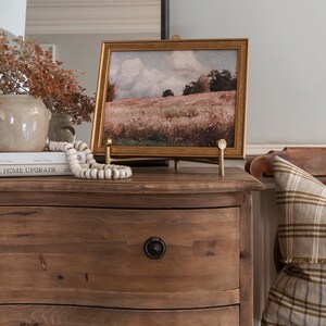 Puede incluir: Una c&oacute;moda de madera con dos cajones y un caballete dorado que sostiene una pintura de paisaje enmarcada. La pintura representa un campo con &aacute;rboles y un cielo nublado. La c&oacute;moda tiene un frente curvo y un acabado marr&oacute;n. El caballete es de metal y tiene un acabado dorado.