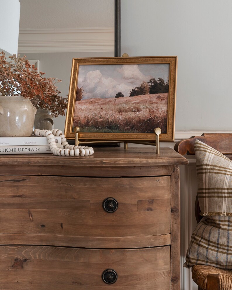 Puede incluir: Una c&oacute;moda de madera con dos cajones y un caballete dorado que sostiene una pintura de paisaje enmarcada. La pintura representa un campo con &aacute;rboles y un cielo nublado. La c&oacute;moda tiene un frente curvo y un acabado marr&oacute;n. El caballete es de metal y tiene un acabado dorado.