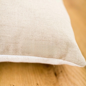 May include: Close-up of a beige linen pillow with a white piping detail. The pillow is lying on a wooden surface.