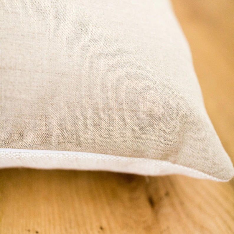 May include: Close-up of a beige linen pillow with a white piping detail. The pillow is lying on a wooden surface.