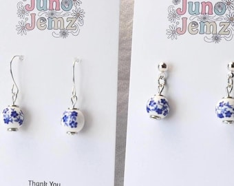 Könnte beinhalten: Zwei Paar Ohrringe auf weißen Präsentationskarten mit der Aufschrift Juno Jemz. Ein Paar besteht aus runden weißen Perlen mit blauem Blumenmuster an silberfarbenen Haken. Das andere Paar hat passende Perlen an silberfarbenen Steckern.