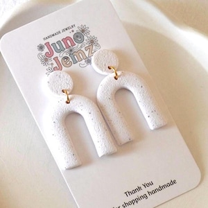 Peut inclure: Boucles d'oreilles pendantes en forme d'arche blanche avec une texture mouchetée, fixées sur une carte de présentation blanche portant le texte "HANDMADE JEWELRY" et "juno jemz" avec des motifs floraux, et "Thank You for shopping handmade".
