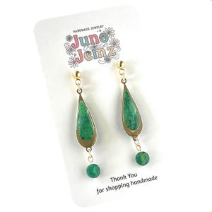 Peut inclure: Boucles d'oreilles pendantes avec une monture dorée en forme de goutte et une incrustation verte texturée, suspendues à un clou doré. Une petite perle verte est fixée à la base de chaque goutte. La carte présente le texte Juno Jemz et Thank You for shopping handmade.