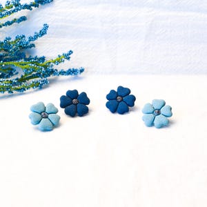 May include: Four flower-shaped stud earrings in varying shades of blue, ranging from light sky blue to deep navy. Each earring features five rounded petals and a small, textured silver-toned centre. The earrings are arranged in a horizontal row.