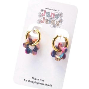 May include: A pair of gold hoop earrings with flower-shaped pendants. The pendants are a mix of pink, purple, blue, and gold. The earrings are displayed on a white card with the text "Juno Jemz" and "Thank You for shopping handmade."