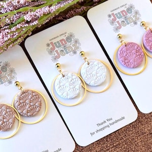 Puede incluir: Pendientes Juno Jemz hechos a mano. Cada par presenta un colgante circular texturizado en blanco, rosa y marrón, suspendido dentro de un aro dorado. Los pendientes se exhiben en tarjetas con el nombre de la marca y el texto "Thank You for shopping handmade."