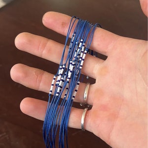Op de afbeelding: Een bundel blauwe en witte kralenarmbanden. De armbanden zijn gemaakt van dun blauw koord en witte kralen. Het koord is aan het einde vastgemaakt met een kleine zilveren kraal.